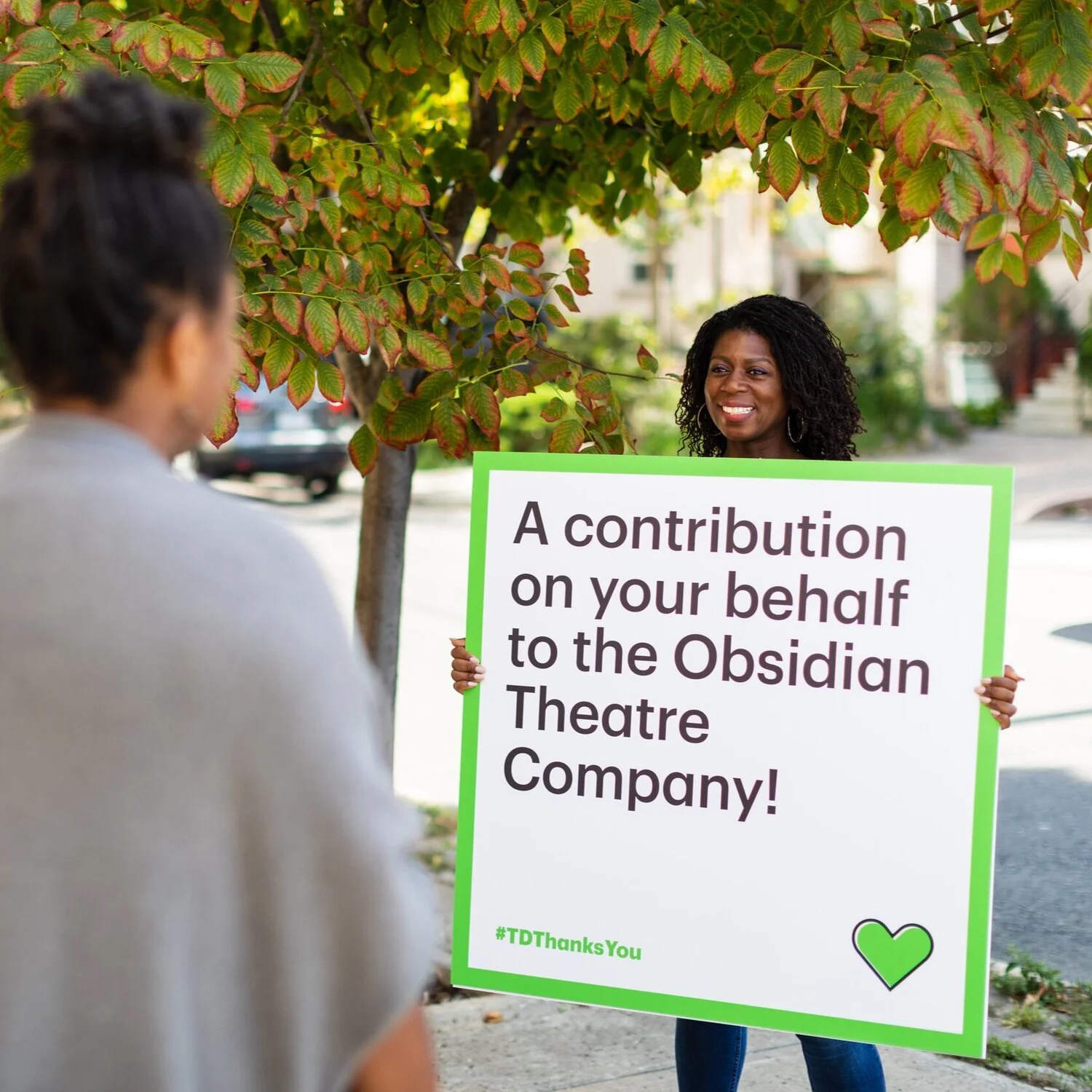 Woman holding a sign for the Obsidian Theatre Company on a sidewalk beside a leafy tree.